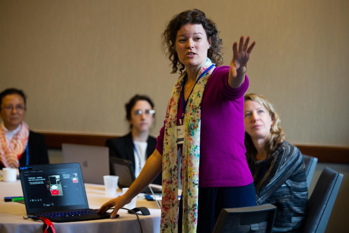 woman speaking to people at a table with a laptop and arm outstretched