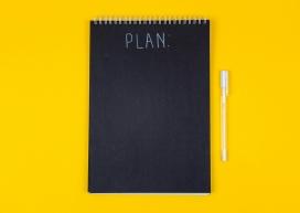 Image contains a black notebook with the word "plan" on it in white against a yellow background