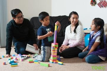 A Latinx family of four (left to right: father, elementary school-aged son, mother, older elementary school-aged daughter) in a living room. The son is playing with building blocks.