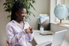 a Black woman wearing a lilac shirt putting her index and middle finger on one hand over her index and middle finger on her other hand while facing a computer